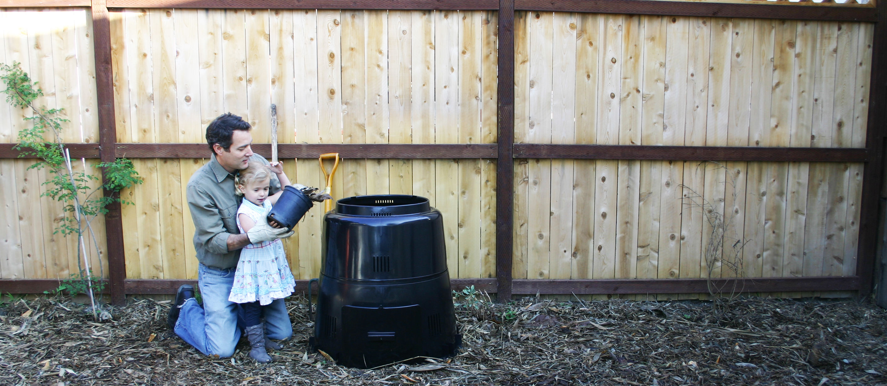 A man helping a young girl deposit the contents of a small black planter into a standing black plastic composter.