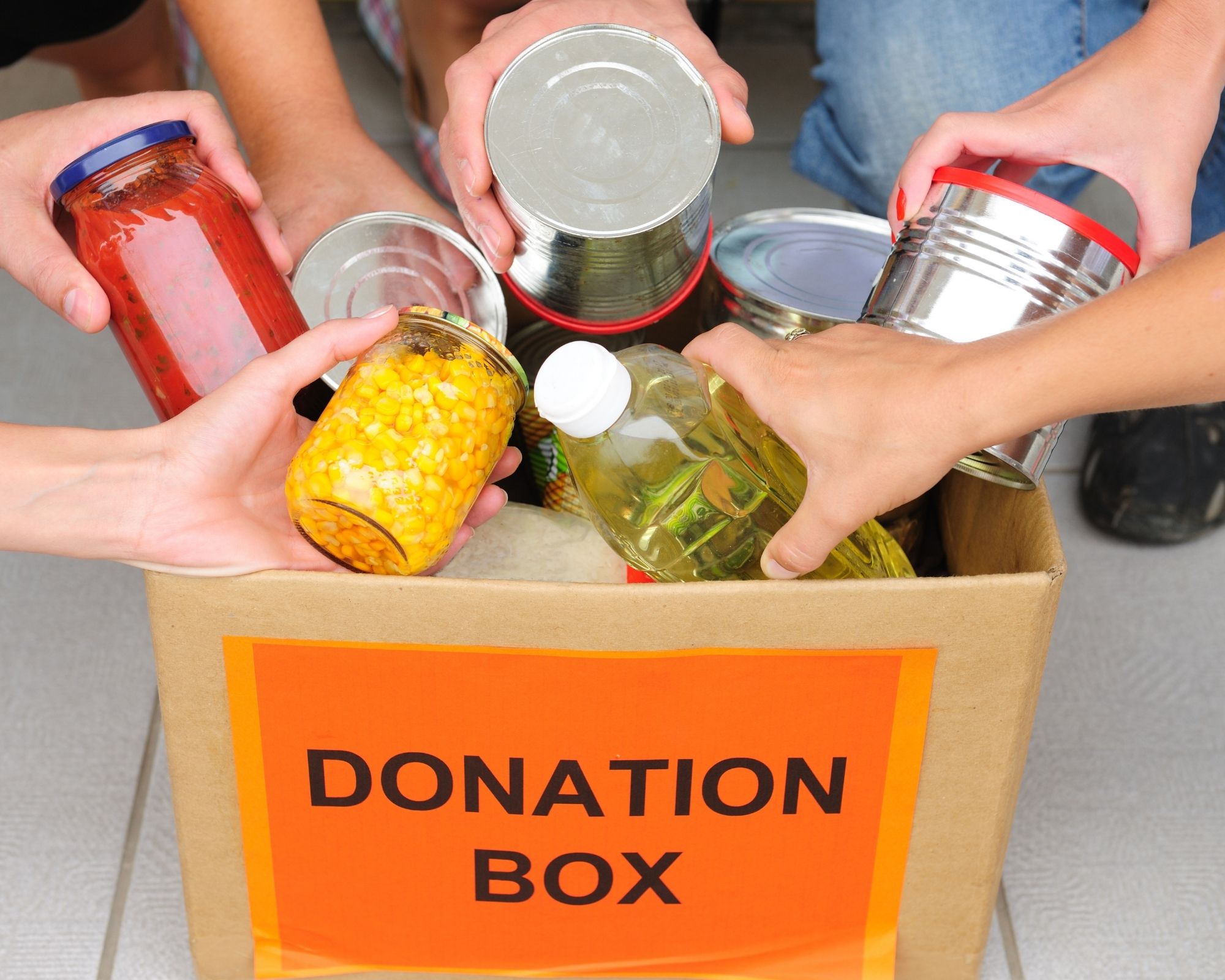 Six hands place various containers of food into a cardboard box labelled "DONATION BOX".