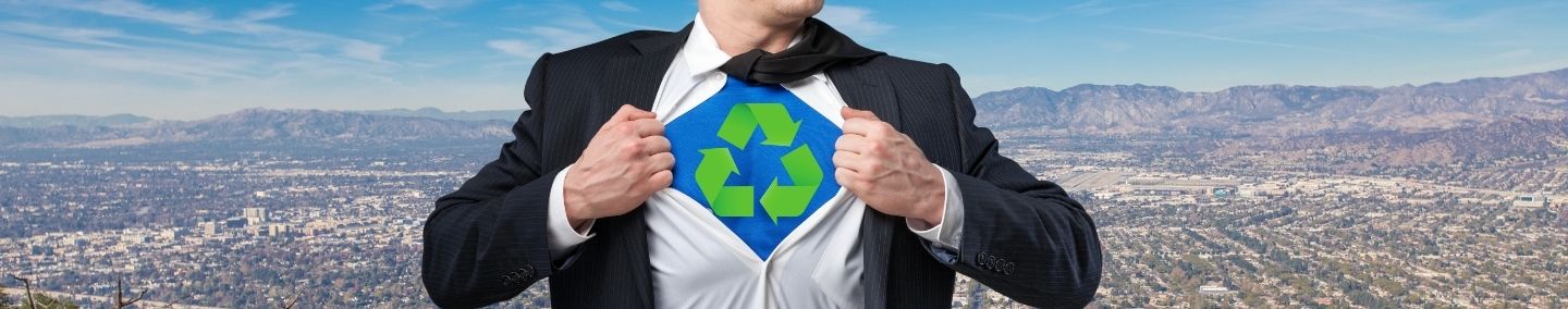 A closeup of a man wearing a suit pulling open his dress shirt to reveal a blue spandex suit and a green recycling logo; a wide shot of the City of Burbank and the mountains is in the background.