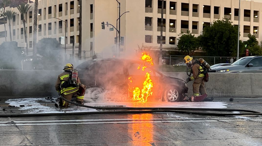 Car on fire in the street, with two firefighters fighting the blaze